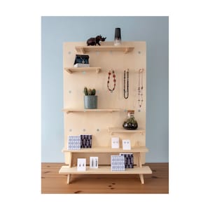 May include: A wooden display stand with multiple shelves, showcasing various jewellery items, a small potted cactus, and decorative objects. The stand is made of light-coloured wood and has a pegboard design. The background is a light blue wall.