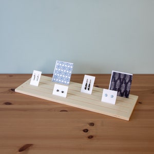 Pu&ograve; includere: Un espositore in legno con cinque fessure per esporre orecchini e carte. L'espositore &egrave; in legno chiaro e ha una finitura naturale. Le carte hanno diversi motivi, tra cui disegni geometrici e floreali. Gli orecchini sono argentati e neri.