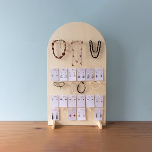 May include: A wooden jewellery display stand with a rounded top. The stand has multiple rows of hooks and cards for displaying necklaces, earrings, and bracelets. The stand is made of light wood and has a natural finish.