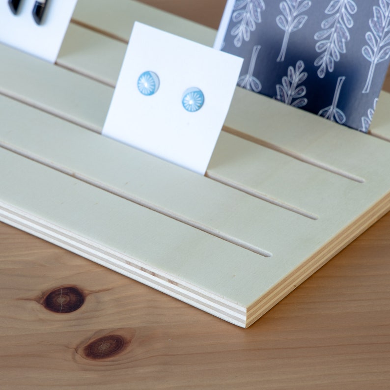 Pu&ograve; includere: Un espositore in legno bianco con fessure per contenere piccoli oggetti, come gioielli o carte. L'espositore ha una venatura del legno chiara ed &egrave; realizzato in compensato. Due piccoli orecchini blu sono esposti su una carta bianca.