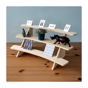 May include: A three-tiered wooden display stand with a curved design. The stand features two cards with geometric patterns, a small succulent plant in a pot, a small figurine of a meditating person, a small figurine of a penguin, and a small wooden rhinoceros figurine.