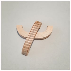 Può includere: Due pezzi di compensato marrone chiaro sono sagomati in una forma curva aperta alle estremità e si incrociano. I pezzi sono uniti al centro.