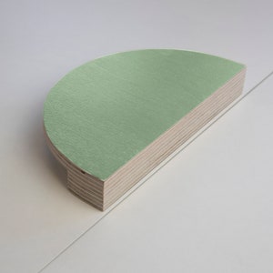 Puede incluir: Un tirador de armario de madera en forma de semicírculo con una superficie pintada de color verde claro. El tirador está hecho de madera en capas, con un borde de madera natural. El tirador del armario es de diseño moderno.