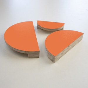 May include: Three orange, semi-circular wooden objects. The objects have a bright orange painted surface and a natural wood edge. The objects are arranged on a white surface. The objects appear to be decorative or functional.
