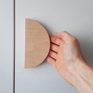 May include: A light wood half-circle cabinet door pull. The pull is mounted on a white cabinet door.