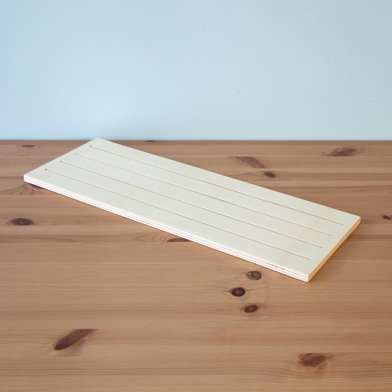 Pu&ograve; includere: Un asse di legno bianco con quattro scanalature parallele che corrono per tutta la sua lunghezza. L'asse &egrave; appoggiato su una superficie di legno marrone chiaro.
