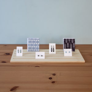 Pu&ograve; includere: Un espositore in legno con cinque fessure per esporre orecchini e carte. L'espositore &egrave; in legno chiaro e ha una finitura naturale. Ci sono due paia di orecchini, una carta con un motivo geometrico e una carta con un motivo ad albero.