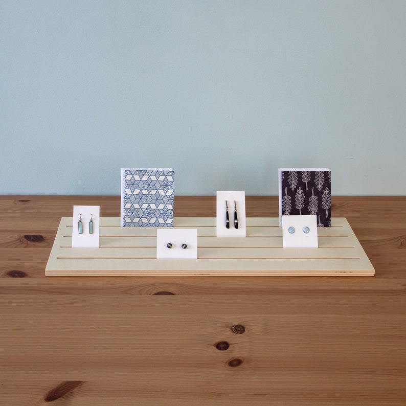Pu&ograve; includere: Un espositore in legno con cinque fessure per esporre orecchini e carte. L'espositore &egrave; in legno chiaro e ha una finitura naturale. Ci sono due paia di orecchini, una carta con un motivo geometrico e una carta con un motivo ad albero.