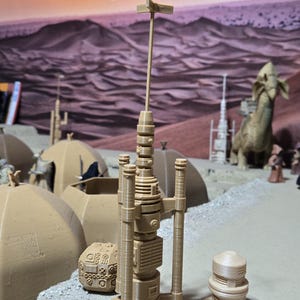 May include: A beige 3D printed model of a Star Wars themed communications tower with a tall antenna. The tower is on a sandy surface with a smaller beige 3D printed model of a dome shaped structure.