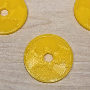 May include: Yellow, circular coasters with a bat symbol and a hexagonal hole in the center. The coasters have a ridged texture and are arranged on a light wood surface.