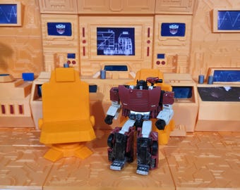 May include: A red and white robot toy with a black chest plate and black legs. The robot is standing on a brown and orange playset with a yellow chair.