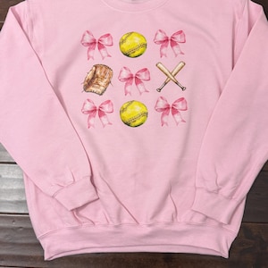 K&ouml;nnte beinhalten: Rosafarbener Pullover mit Softball-Motiv. Das Design zeigt zwei Softb&auml;lle, einen Handschuh, zwei Schl&auml;ger und rosafarbene Schleifen.
