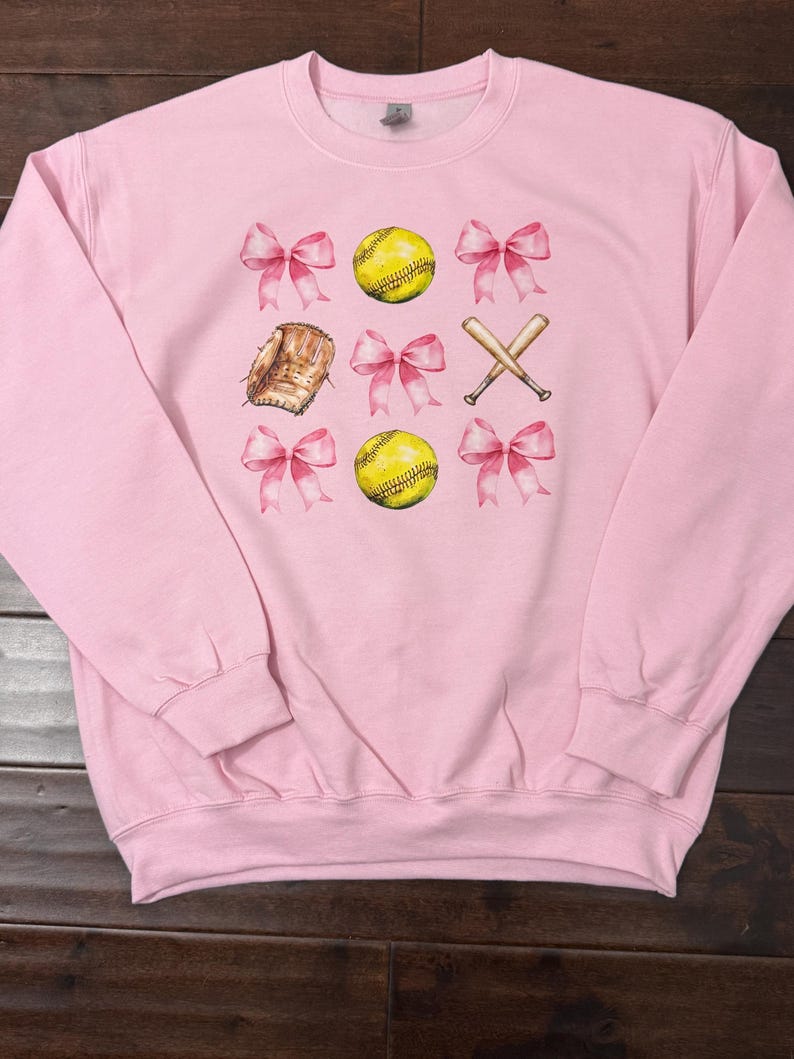 K&ouml;nnte beinhalten: Rosafarbener Pullover mit Softball-Motiv. Das Design zeigt zwei Softb&auml;lle, einen Handschuh, zwei Schl&auml;ger und rosafarbene Schleifen.