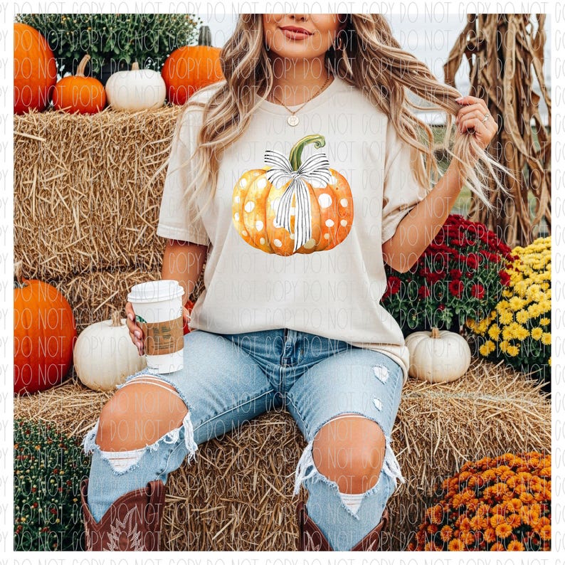 May include: A beige t-shirt featuring a watercolor pumpkin design with a white bow and polka dots. The pumpkin is orange and yellow. The person is wearing blue jeans with ripped knees. The background includes hay bales and pumpkins.