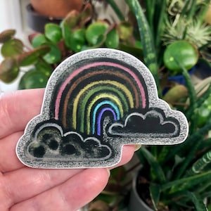 Puede incluir: Una pegatina con un diseño de arcoíris y nubes oscuras. El arcoíris tiene rayas rosas, marrones, amarillas, verdes y azules. La pegatina se sostiene en una mano, con plantas en el fondo.