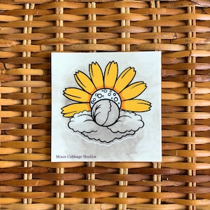 Puede incluir: Adhesivo cuadrado con un diseño caprichoso de un girasol amarillo, una nube blanca y gris y un objeto blanco en el centro. El texto "Moon Cabbage Studios" está impreso en la parte inferior.