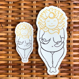 Puede incluir: Dos pegatinas blancas con un diseño de diosa solar. La pegatina más grande presenta una cara de sol sonriente con un halo y una figura femenina que sostiene sus senos. La pegatina más pequeña tiene el mismo diseño. Las pegatinas están delineadas en negro y amarillo.