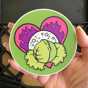 Puede incluir: Una pegatina redonda con un fondo verde que presenta un diseño de dibujos animados. El diseño incluye una col verde claro, un círculo blanco con puntos y un diseño de llama en forma de corazón púrpura y rosa. La pegatina se sostiene en una mano.