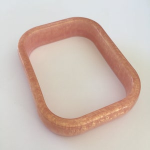 May include: A square-shaped bracelet in a light peach color with a subtle, speckled pattern. The bracelet has rounded corners and a smooth, glossy finish. The jewelry piece is displayed on a white surface.