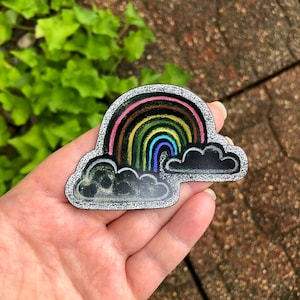 Puede incluir: Un pin colorido con forma de arcoíris y nube. El arcoíris presenta rayas rosas, marrones, amarillas, verdes y azules. Las nubes son negras con un contorno blanco. El pin se sostiene en una mano.