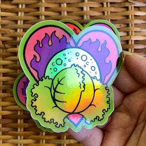 Puede incluir: Un conjunto de pegatinas holográficas en forma de corazón. Las pegatinas presentan una imagen central verde y amarilla con un diseño de hojas, rodeada por un diseño de llama rosa y púrpura. Las pegatinas tienen un acabado brillante e iridiscente.
