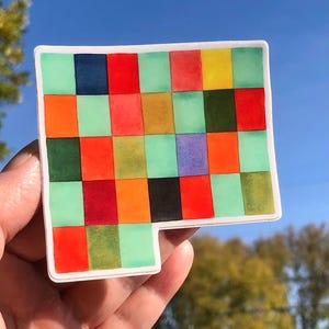 Puede incluir: Una colorida pegatina con un patrón de cuadrícula de cuadrados en varios colores, incluyendo rojo, naranja, verde y amarillo. La pegatina tiene un borde blanco y una forma recortada única en un lado. El fondo es un cielo azul claro.