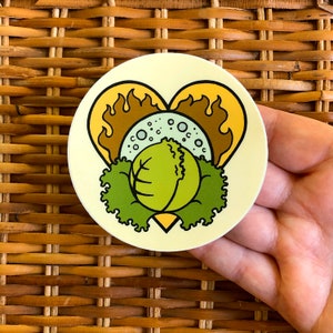 Puede incluir: Una pegatina redonda con un diseño en forma de corazón que presenta una col verde, lechuga y una llama marrón y amarilla. La pegatina tiene un fondo amarillo claro y está sujeta en una mano.
