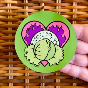 Puede incluir: Una pegatina redonda con un fondo verde que presenta un diseño de col. La col está rodeada por una forma de corazón con llamas moradas y acentos rosas. La pegatina está sujeta por una mano.