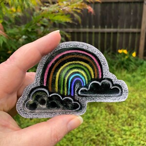 Puede incluir: Una pegatina de arcoíris colorida con un contorno negro y dos nubes negras. El arcoíris presenta rayas rosas, marrones, amarillas, verdes y azules. La pegatina se sostiene en una mano sobre un fondo de hierba verde y una valla de madera.
