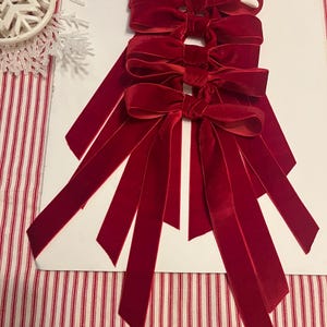 May include: Five deep red velvet bows with long tails are displayed on a white surface. The bows, made of soft velvet, are tied and have long, flowing tails. The bows are a rich, deep red colour, perfect for festive decorations.