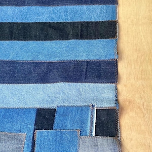 Peut inclure: Textile patchwork en denim bleu de différentes nuances. Le motif présente des pièces rectangulaires et carrées cousues ensemble, créant une surface texturée. Les bords sont finis avec des coutures visibles.