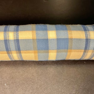 May include: A long, cylindrical pillow with a blue and tan plaid pattern. The pillow's fabric has a woven texture, and the plaid design features alternating squares and stripes of blue and tan. The pillow is likely used for decorative or support purposes.
