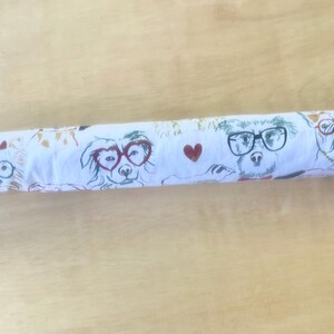 May include: A white fabric roll featuring a repeating pattern of various dog breeds wearing glasses, including heart-shaped and round frames, interspersed with small red hearts. The dogs are illustrated in a sketch style with black and red ink details.