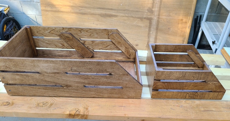 May include: Two wooden crates, one larger than the other, both with a brown stain finish. The larger crate has a slanted front and a handle on the side. The smaller crate has a rectangular shape and a handle on the side.