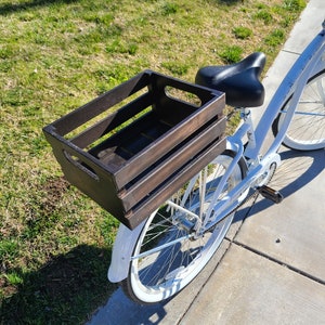 Pode incluir: Uma bicicleta branca com uma caixa de madeira marrom presa ao porta-bagagens traseiro. A bicicleta tem um assento preto e pneus brancos.