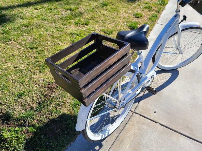 Pode incluir: Uma bicicleta branca com uma caixa de madeira marrom presa ao porta-bagagens traseiro. A bicicleta tem um assento preto e pneus brancos.