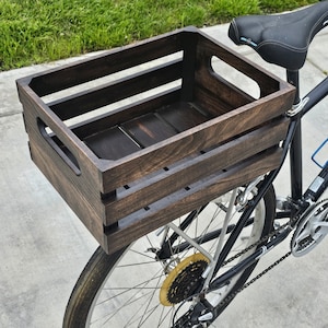 Pode incluir: Uma caixa de madeira marrom escura presa ao porta-bagagens traseiro de uma bicicleta preta. A caixa tem uma al&ccedil;a de cada lado e &eacute; feita de madeira resistente.