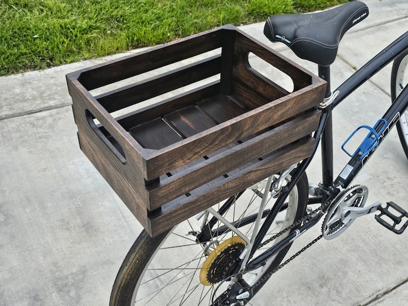 Pode incluir: Uma caixa de madeira marrom escura presa ao porta-bagagens traseiro de uma bicicleta preta. A caixa tem uma al&ccedil;a de cada lado e &eacute; feita de madeira resistente.