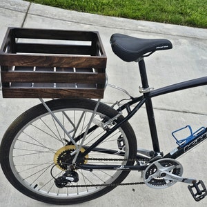 Pode incluir: Uma bicicleta preta com uma caixa de madeira presa ao porta-bagagens traseiro. A caixa &eacute; tingida de uma cor marrom escura e tem uma al&ccedil;a de metal. A bicicleta tem um assento preto e um porta-garrafas.