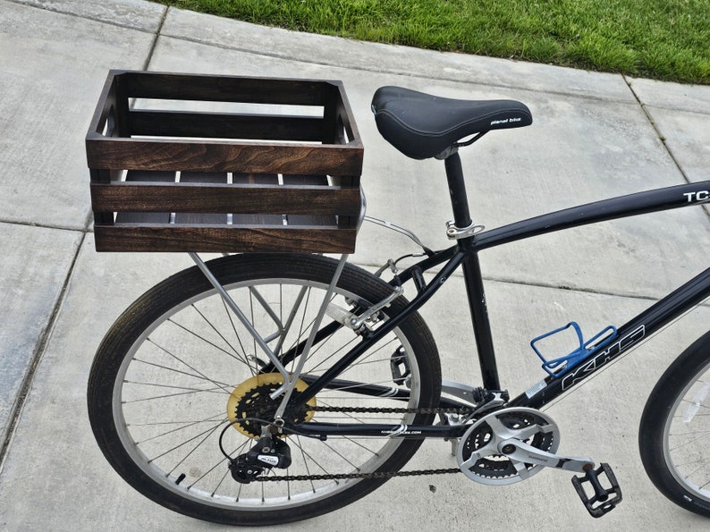 Pode incluir: Uma bicicleta preta com uma caixa de madeira presa ao porta-bagagens traseiro. A caixa &eacute; tingida de uma cor marrom escura e tem uma al&ccedil;a de metal. A bicicleta tem um assento preto e um porta-garrafas.