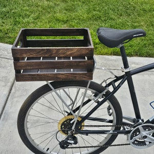 Pode incluir: Uma bicicleta preta com uma caixa de madeira presa ao porta-bagagens traseiro. A caixa &eacute; tingida de uma cor marrom escura e tem uma moldura de metal.