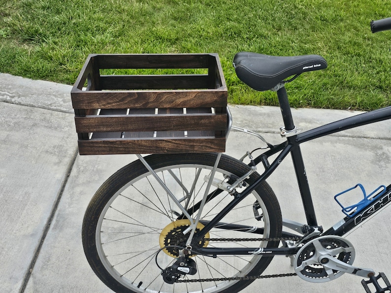 Pode incluir: Uma bicicleta preta com uma caixa de madeira presa ao porta-bagagens traseiro. A caixa &eacute; tingida de uma cor marrom escura e tem uma moldura de metal.