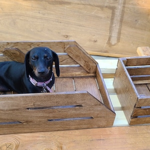 May include: A black dachshund dog sits inside a wooden crate with a light brown stain. The crate has a decorative design with a raised edge. A smaller wooden crate with a similar design is next to the larger crate.