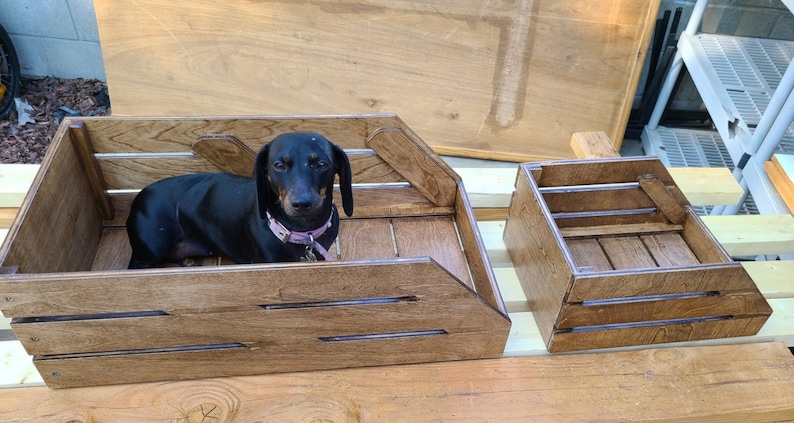 May include: A black dachshund dog sits inside a wooden crate with a light brown stain. The crate has a decorative design with a raised edge. A smaller wooden crate with a similar design is next to the larger crate.