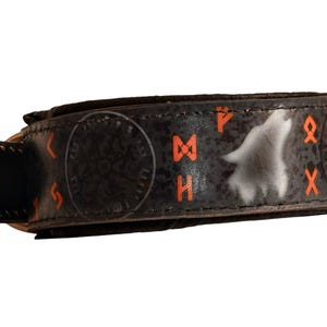 Puede incluir: Brazalete de cuero negro con símbolos rúnicos naranjas y un diseño de lobo aullando. El brazalete tiene un cierre de hebilla y es probablemente un accesorio de moda o cosplay.