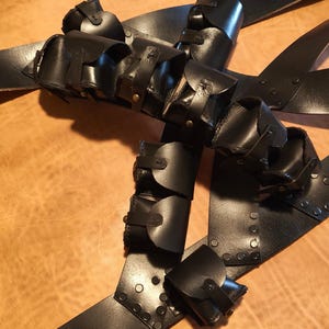 Pode incluir: Arnês de couro preto com vários bolsos e correias. O arnês apresenta pequenos detalhes rebitados e fivelas ajustáveis. Os bolsos são presos com abas e pequenas fivelas. O design sugere um propósito utilitário ou tático.