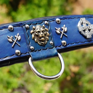May include: Blue leather dog collar with silver Celtic cross, lion head, and axe accents. The collar has silver studs and a silver D-ring.