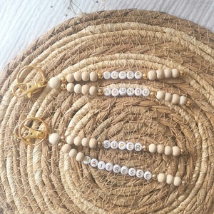 Peut inclure: Deux porte-clés en perles de bois avec des accents dorés. Les perles sont blanches avec des lettres dorées qui épellent "SUPER NOUNOU" et "SUPER MAITRESSE".
