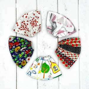 May include: Five fabric doll skirts with different patterns. The skirts are made of cotton and have a variety of colors and designs, including red, white, blue, black, green, and yellow. The skirts are arranged in a circle on a white wooden surface.