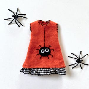 Puede incluir: Un vestido de fieltro naranja con un dise&ntilde;o de ara&ntilde;a negra y un dobladillo a cuadros blanco y negro. Dos ara&ntilde;as de pl&aacute;stico negro se colocan a cada lado del vestido. El vestido tiene costuras negras.